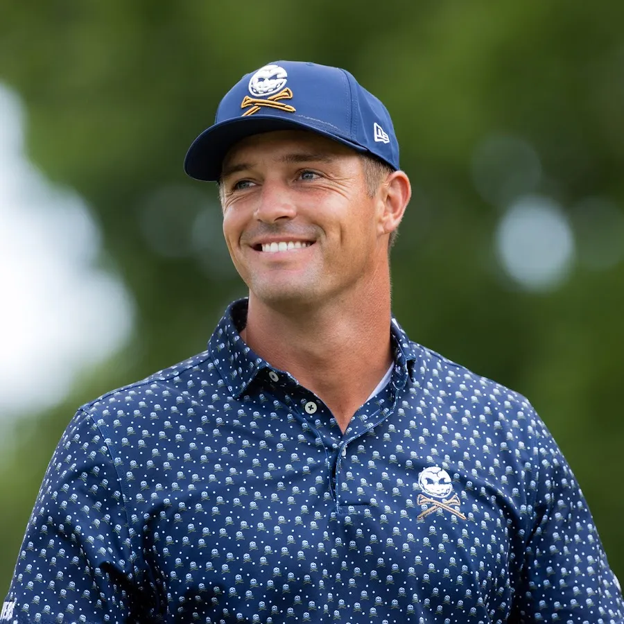 Bryson DeChambeau smiling on a golf course, in a navy patterned polo and cap.